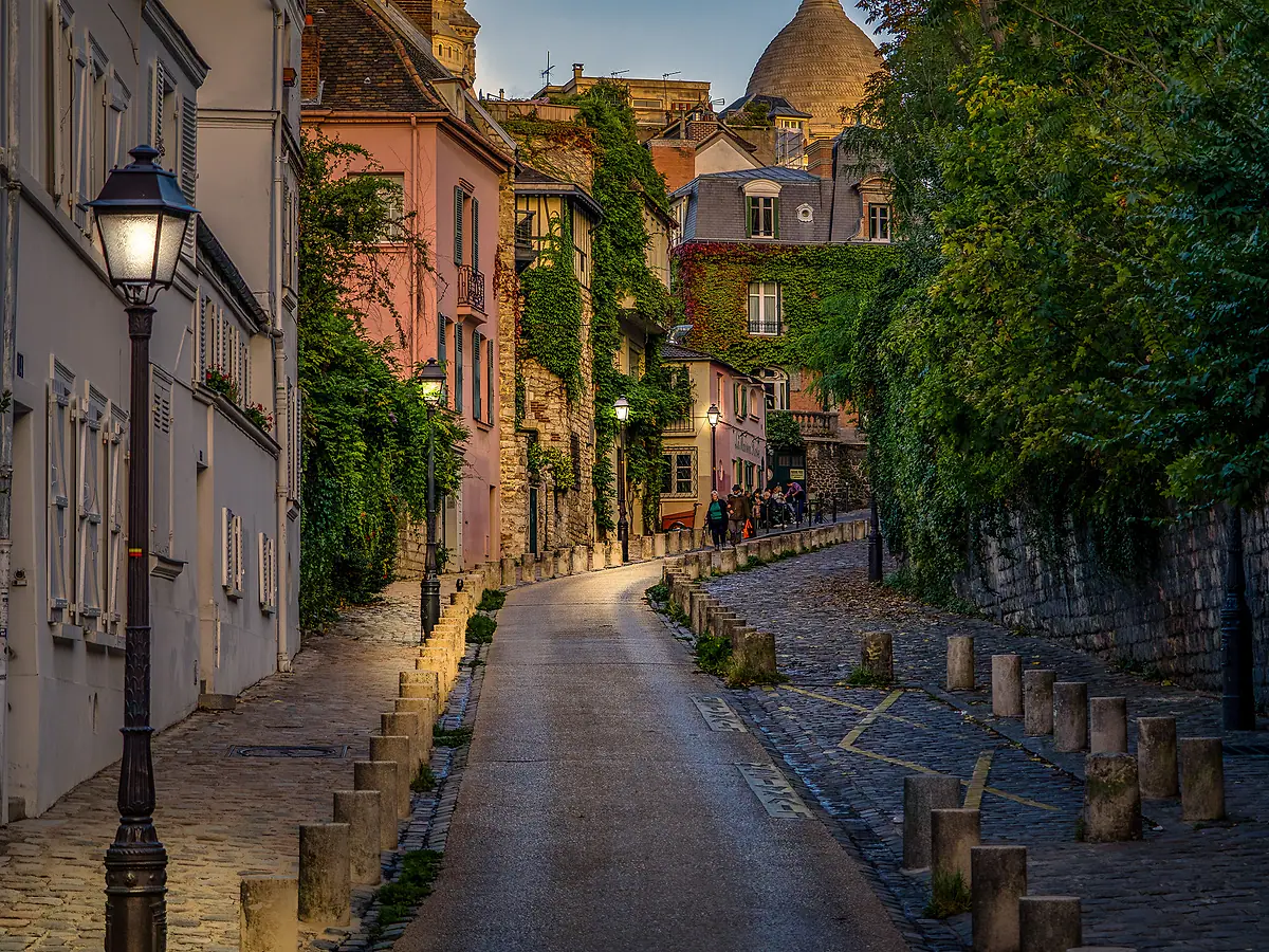 A lamp-lit street in Montmartre, Paris, with the domes of Sacré-Cœur above the rooftops.